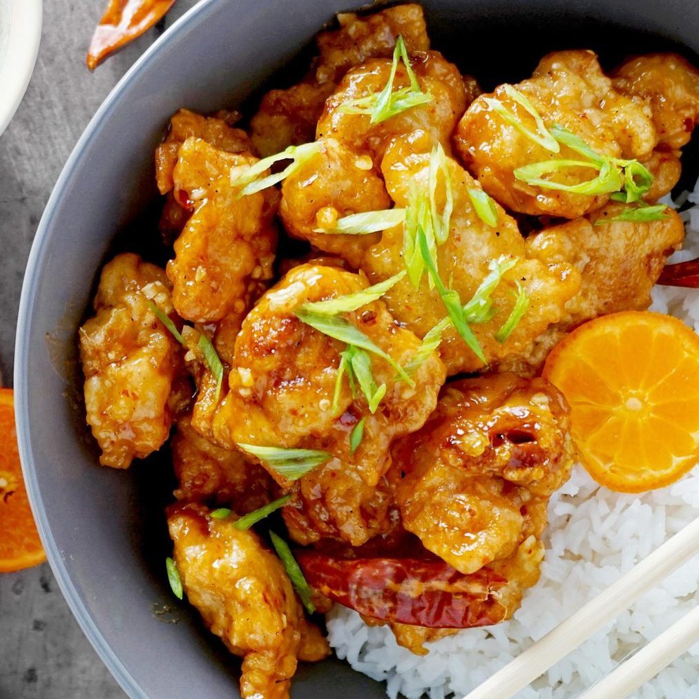 Fried chicken rests on white rice, garnished with green onions, surrounded by orange slices and red peppers in a gray bowl.