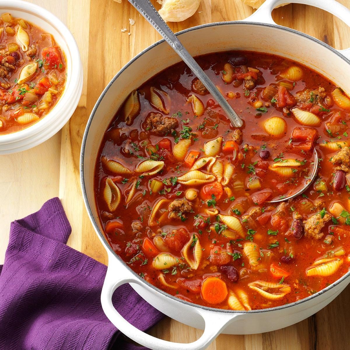 Pot of soup sits on wooden surface; features shell pasta, carrots, beans, and meat in a tomato broth. Purple cloth and bowl nearby.