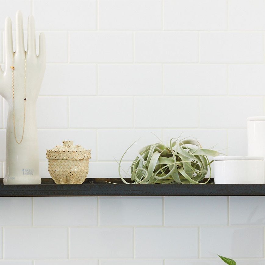 Ceramic hand displays necklace on black shelf; beside it, a woven container sits next to a small plant and white pots, against tiled wall.