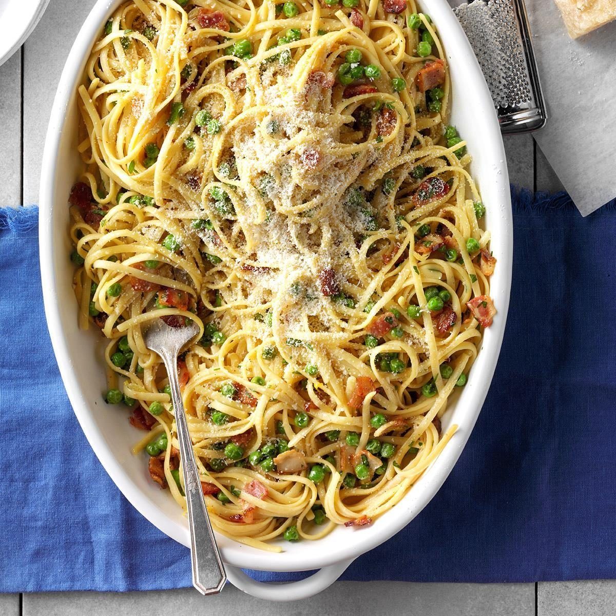 Pasta sits in a white dish, topped with cheese, mixed with peas and bacon; surrounded by a blue cloth on a table.