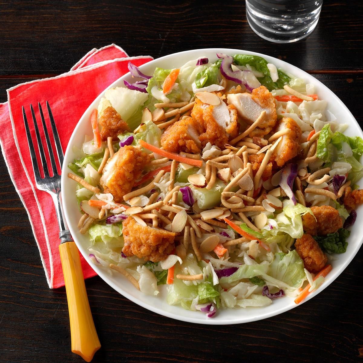 Salad topped with crispy chicken and sliced almonds, resting on a wooden table, accompanied by a fork on a red napkin and a glass of water.