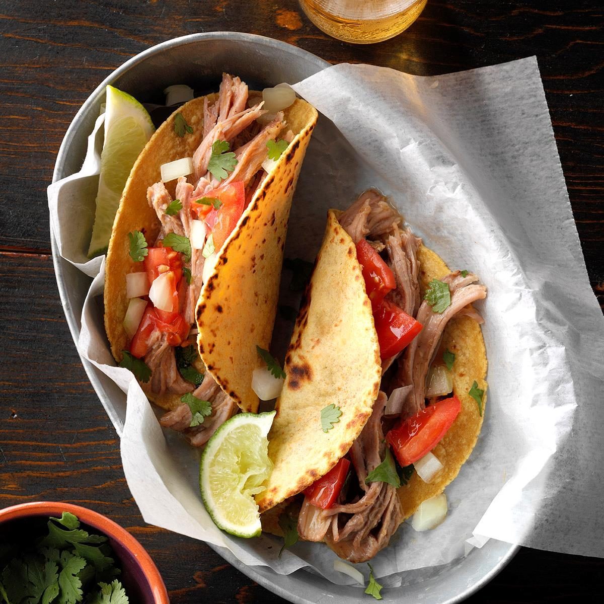 Tacos filled with shredded meat, diced tomatoes, onions, and cilantro rest on parchment paper in a metal dish with lime wedges nearby.