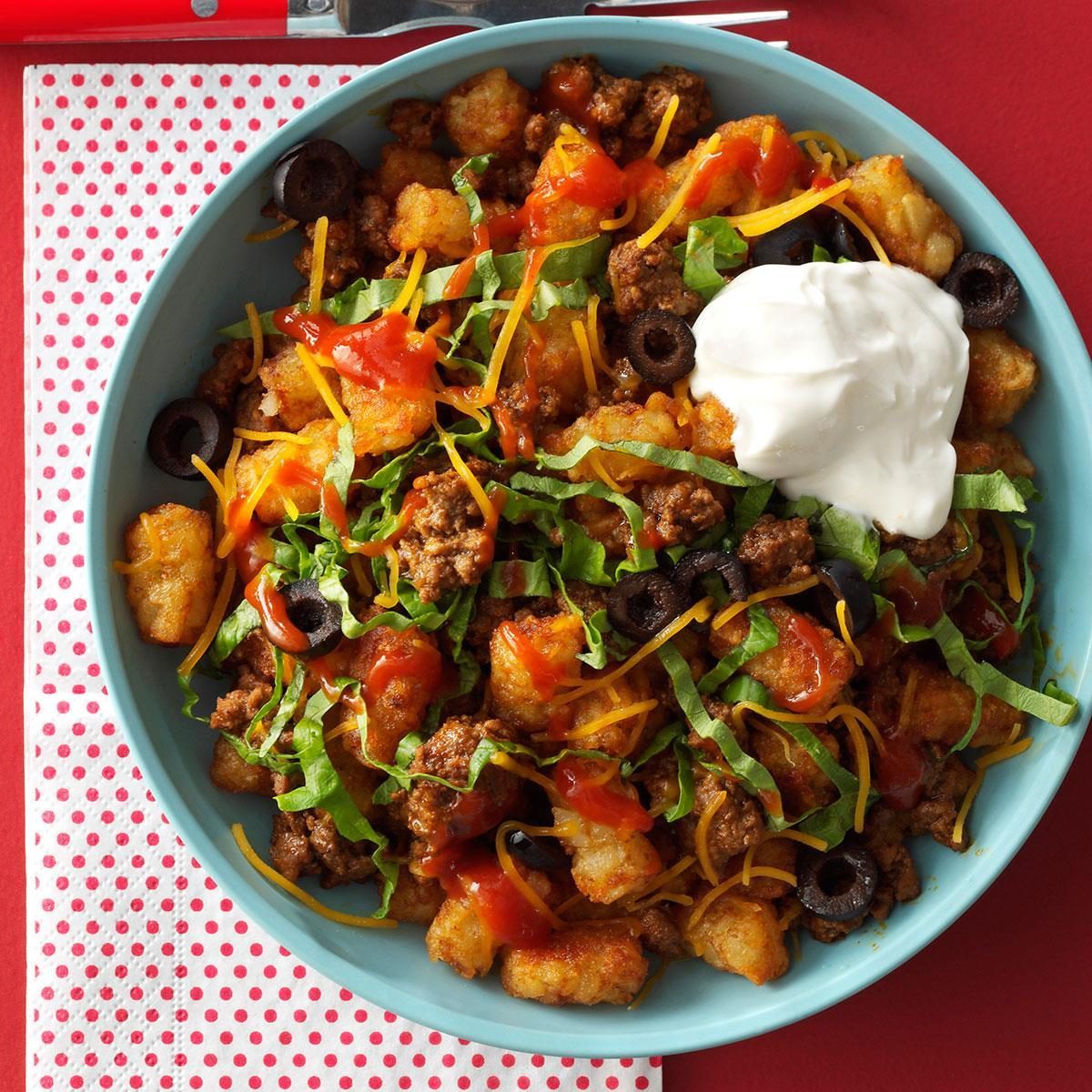 Bowl holds loaded tater tots with ground beef, cheese, lettuce, black olives, salsa, and a dollop of sour cream, on a red background.