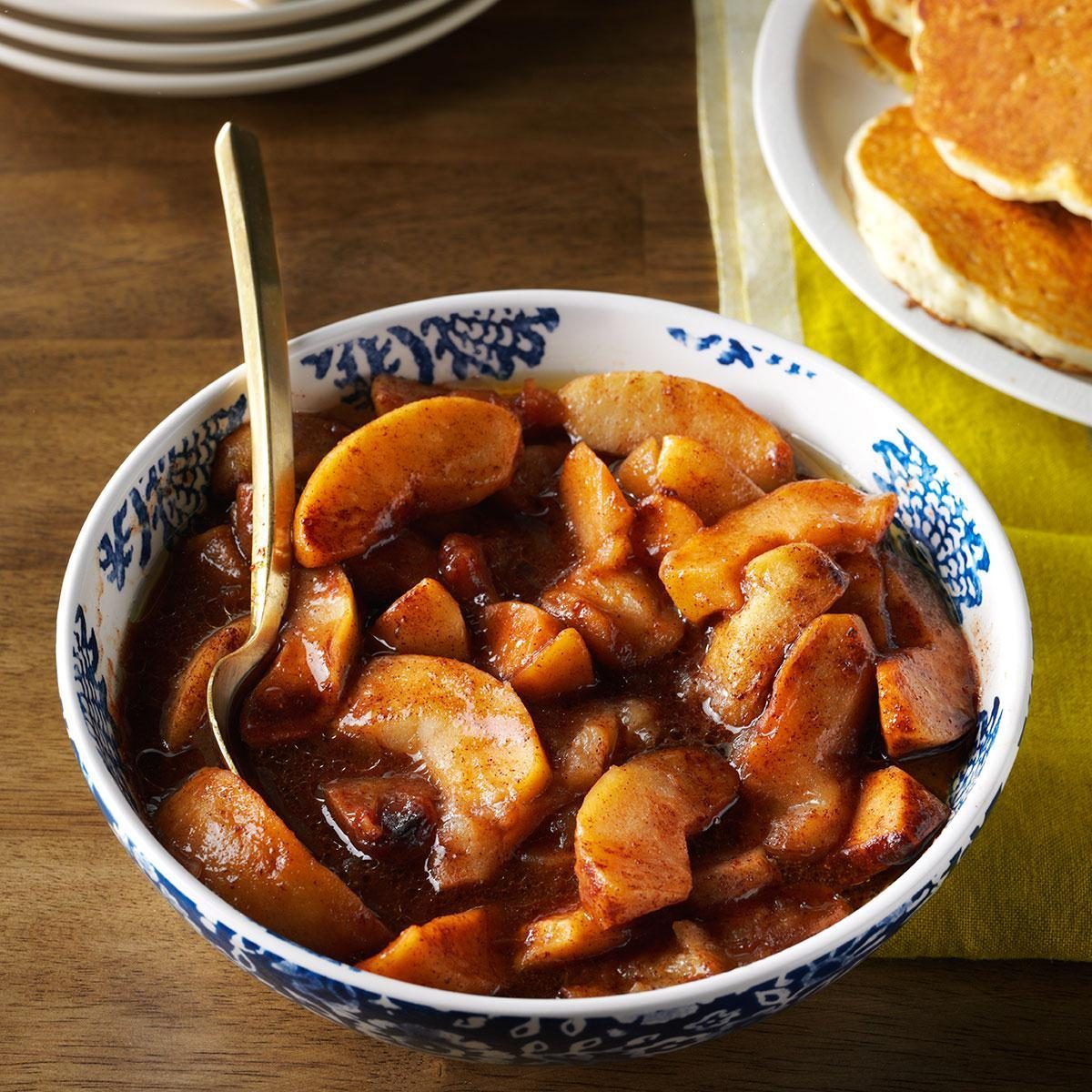 Bowl holding cinnamon-glazed apple slices with a spoon, placed on a wooden table beside a plate of pancakes.