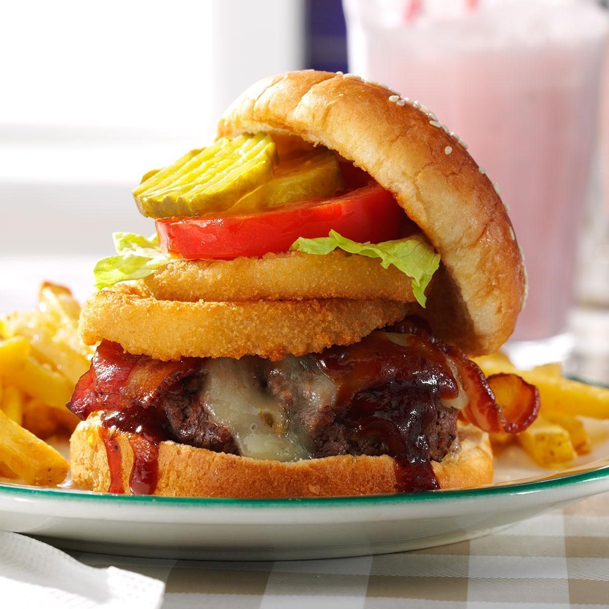 Burger with bacon and onion rings sits on a plate beside fries; milkshake in the blurred background.