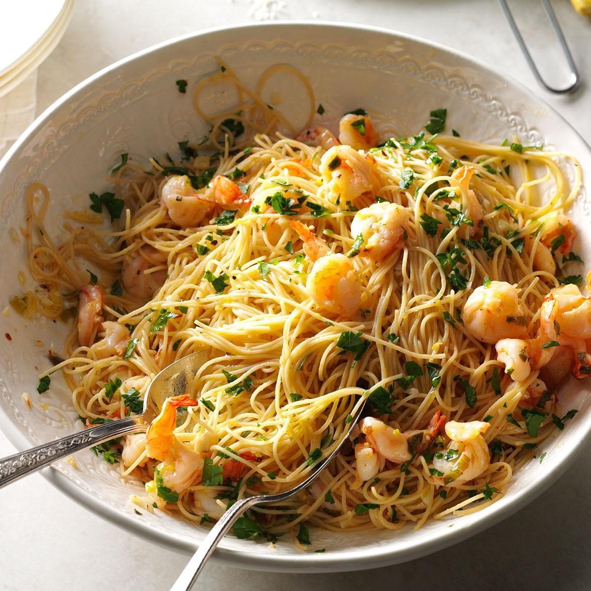 Pasta sits in a large white bowl, mixed with shrimp and sprinkled with chopped parsley, surrounded by a light, airy dining setting.
