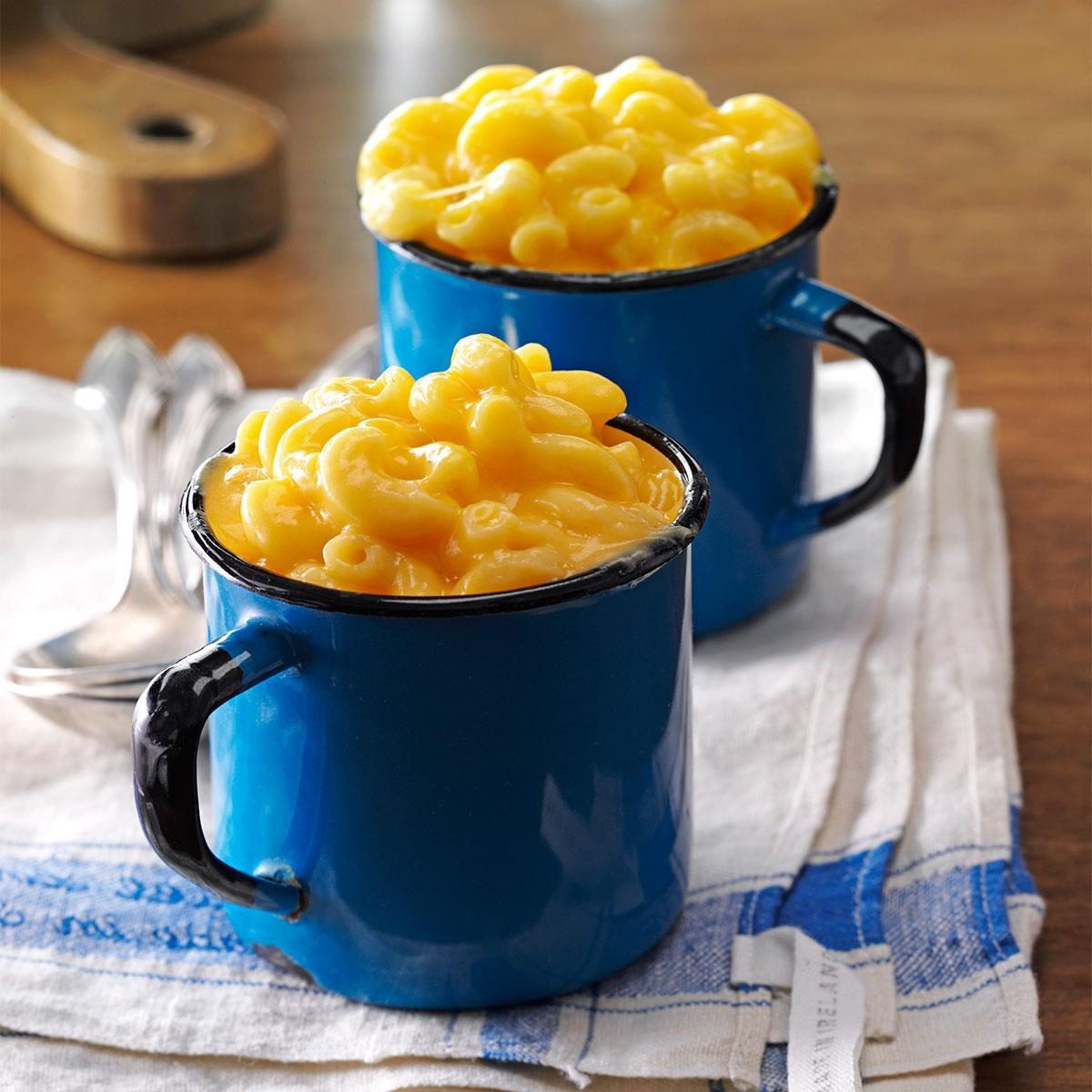 Macaroni and cheese fills blue mugs, resting on a folded cloth with spoons nearby on a wooden table.