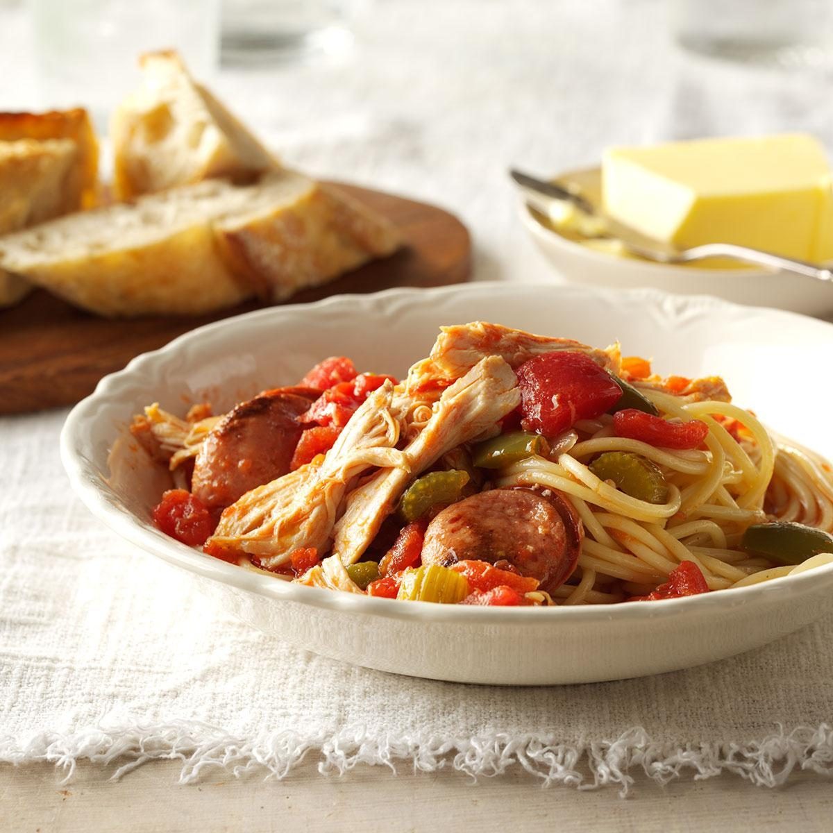 Pasta topped with chicken, sausage, and vegetables sits in a white bowl, surrounded by sliced bread and butter on a tablecloth.