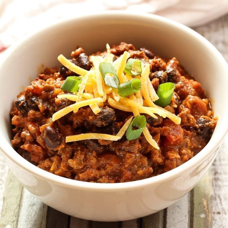 Bowl holds chili, topped with shredded cheese and chopped green onions, resting on a rustic wooden surface.