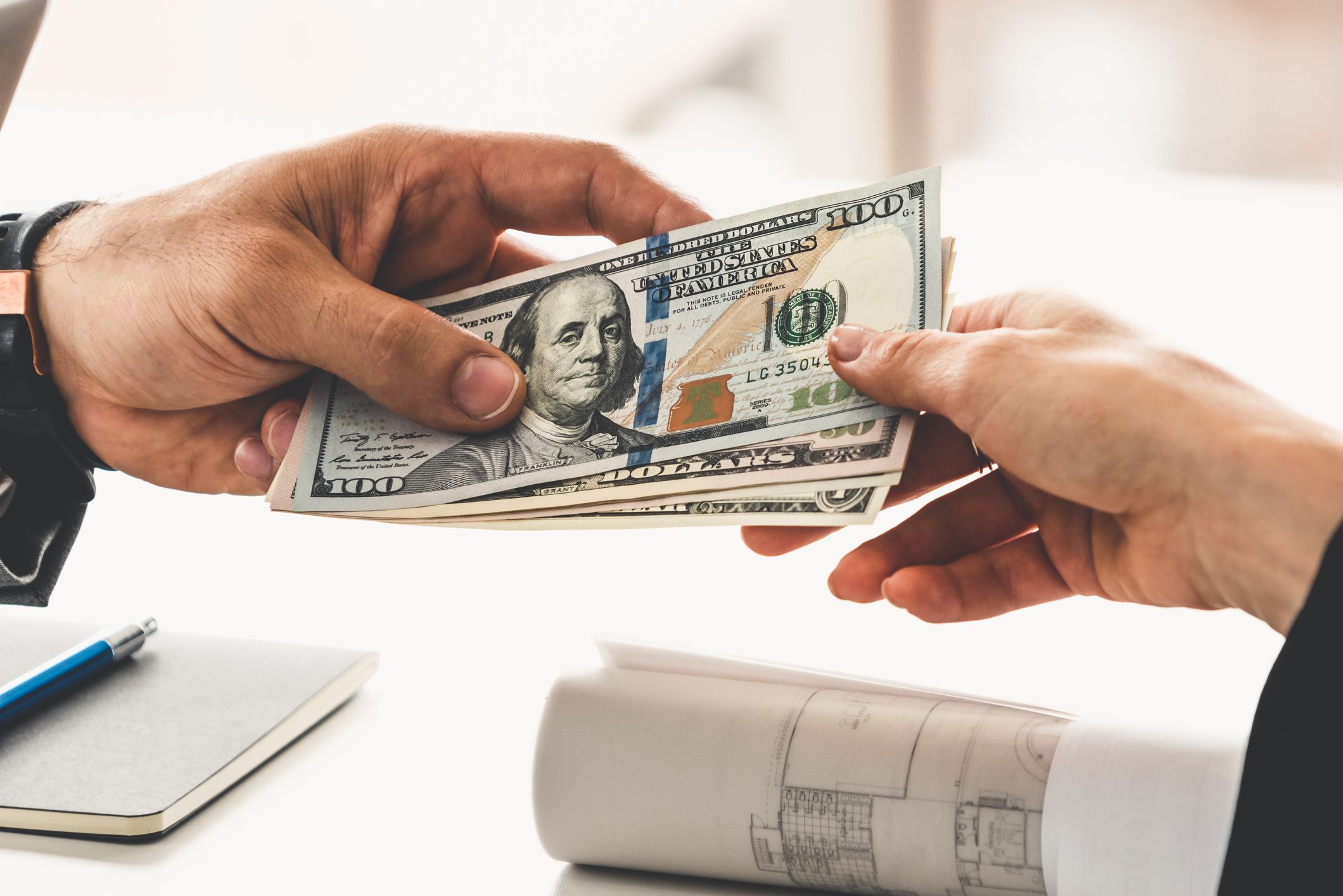 Hands exchange dollar bills over a table with a notebook, pen, and architectural plans.