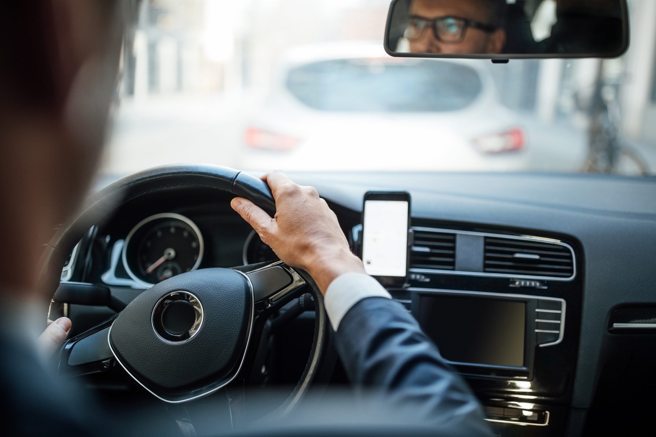 Driver navigates car using steering wheel; smartphone mounted on dashboard, city street in view ahead.