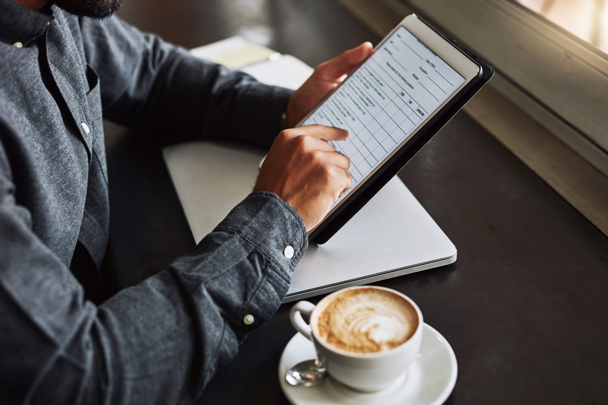 Person holding a tablet, entering information on a form, with a laptop and a cup of cappuccino on a dark table nearby.