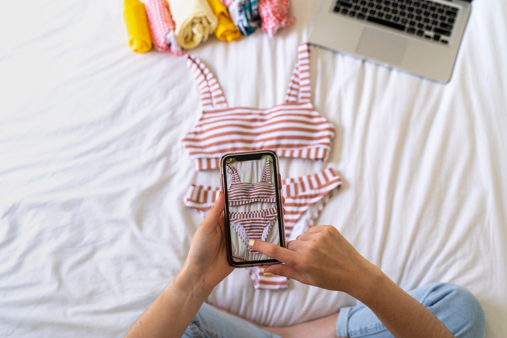 Hands photograph a striped bikini on a smartphone, surrounded by rolled clothes and a laptop on a white bed.