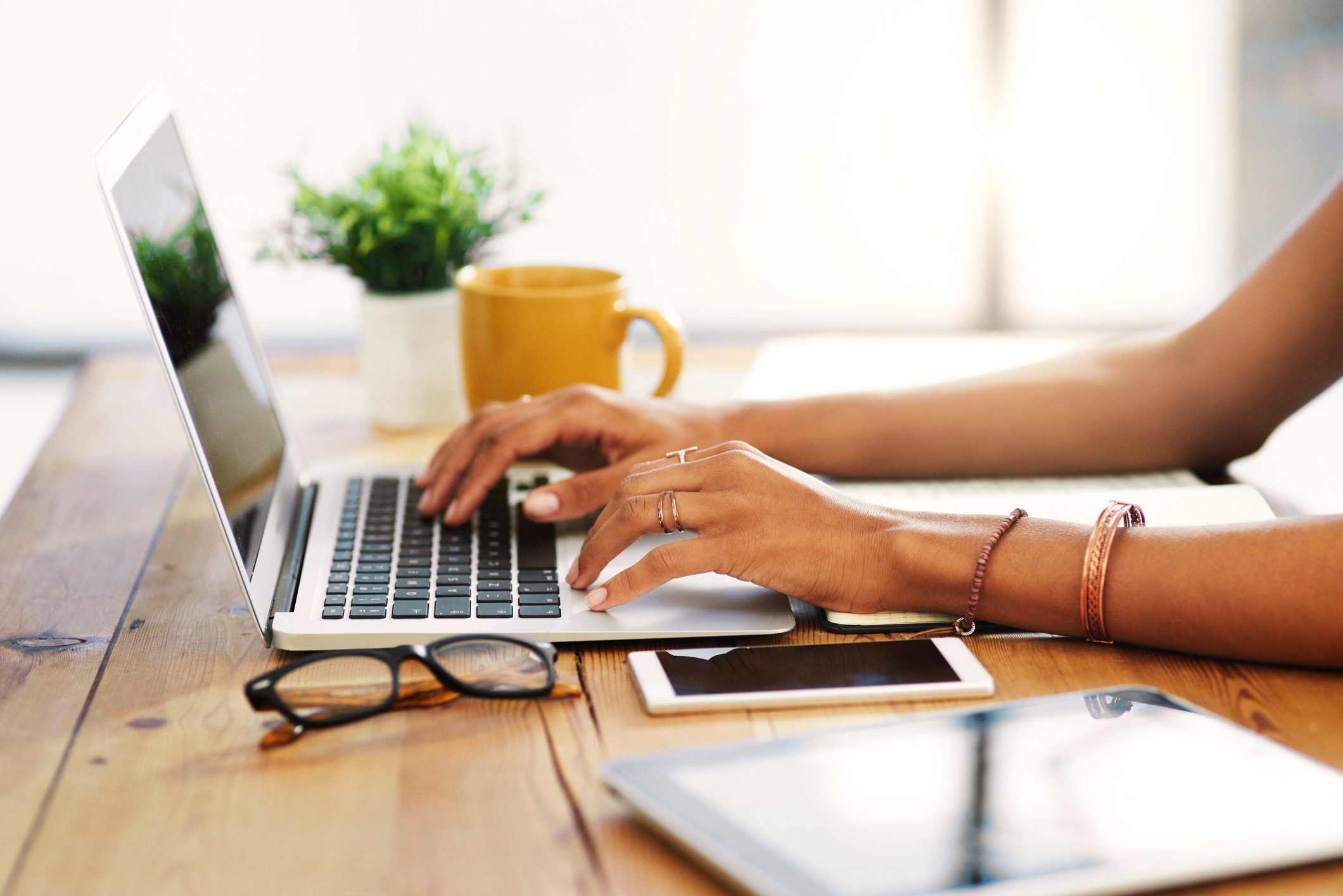 Hands typing on a laptop, sitting on a wooden table with glasses, smartphone, and a yellow mug nearby.