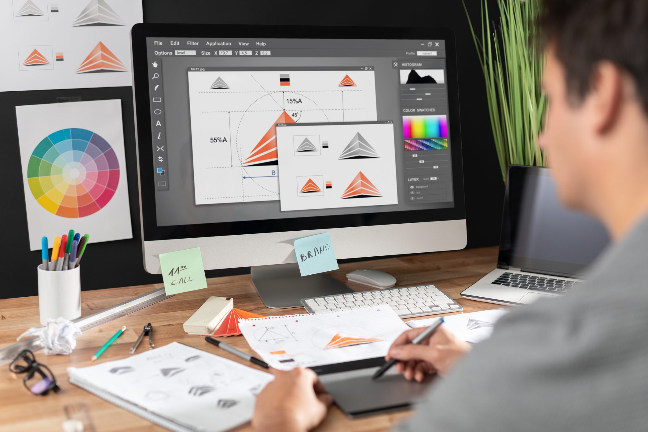 Designer creates geometric graphics on a computer in a workspace with color wheel, sketches, and office supplies. Sticky notes read "11:00 CALL" and "BRAND."