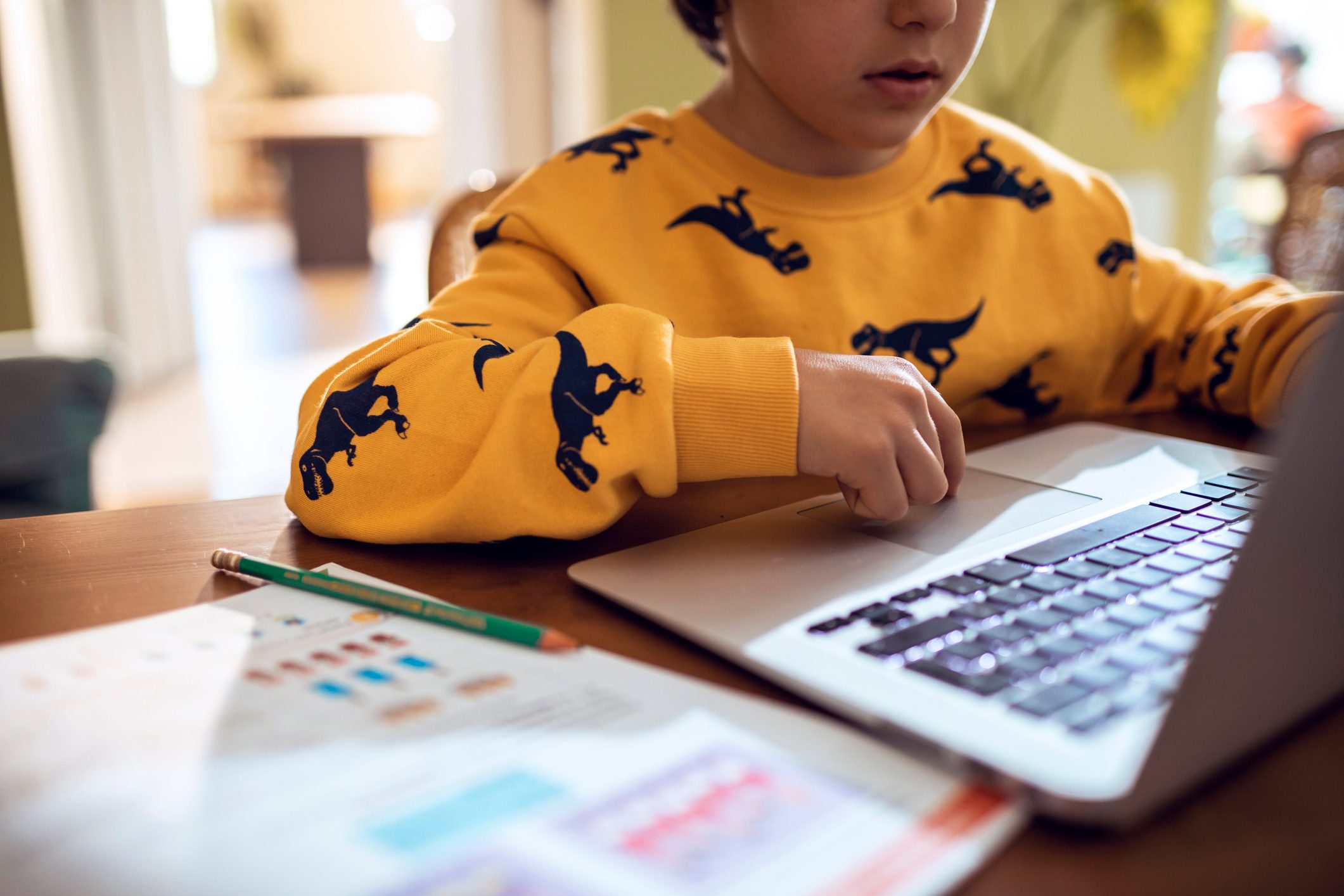 Child uses laptop at wooden table, wearing yellow dinosaur sweater. Nearby, a green pencil and printed worksheets are visible in a home setting.