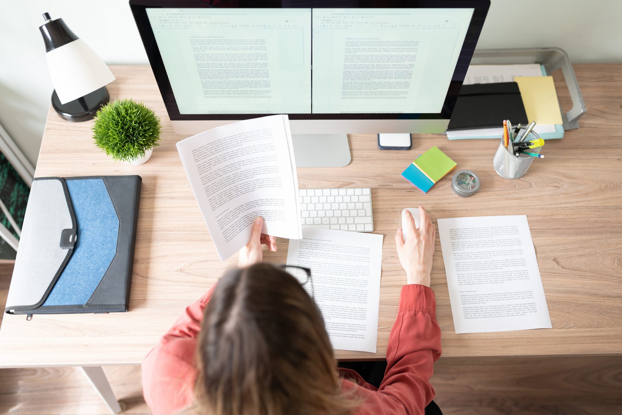 Person reads documents while using a computer in a well-organized office space with stationery and a small plant on the desk.