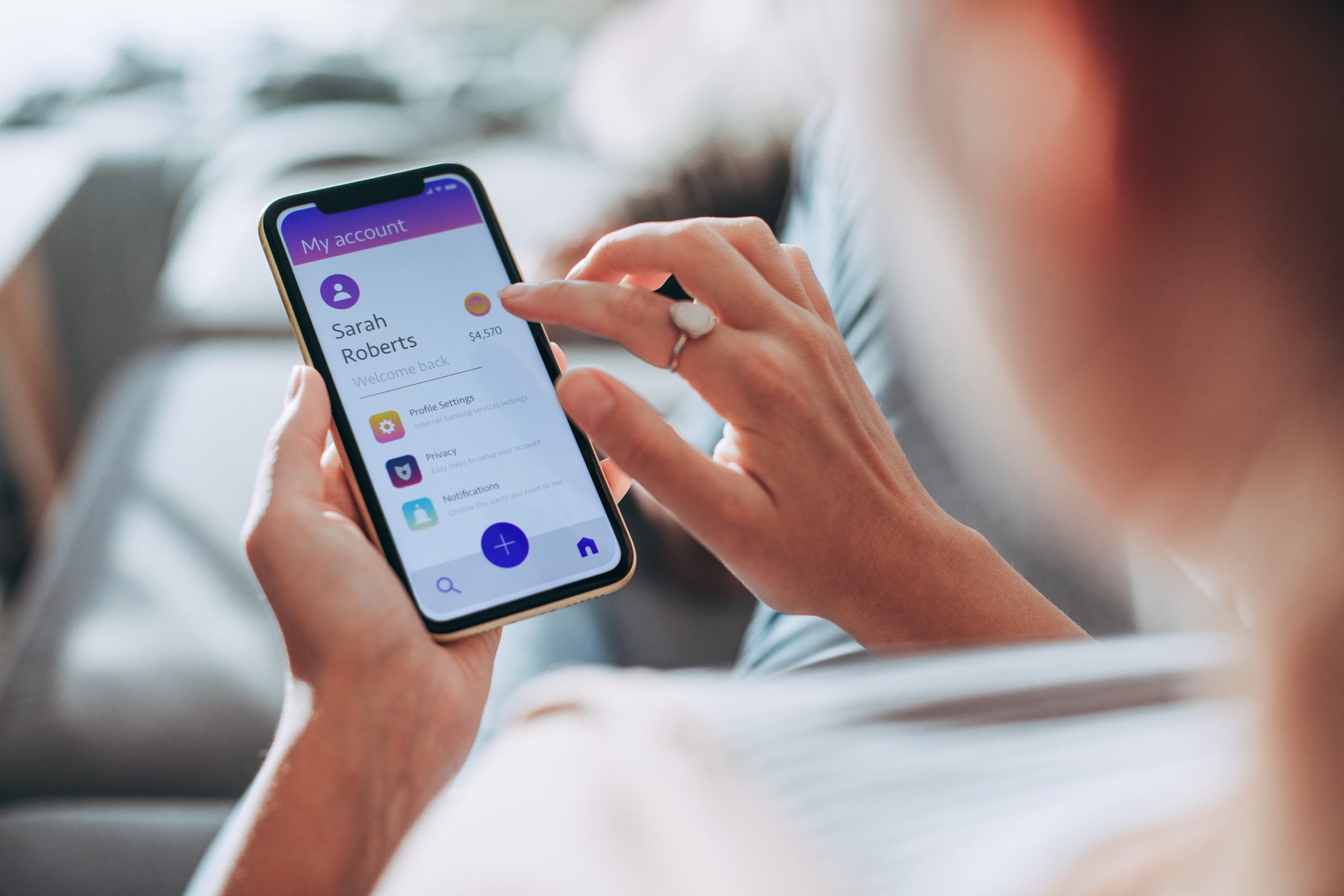 Hand interacting with smartphone, displaying a banking app. Text includes "My account," "Sarah Roberts," and balance "$A 570." Background is a blurred indoor setting.