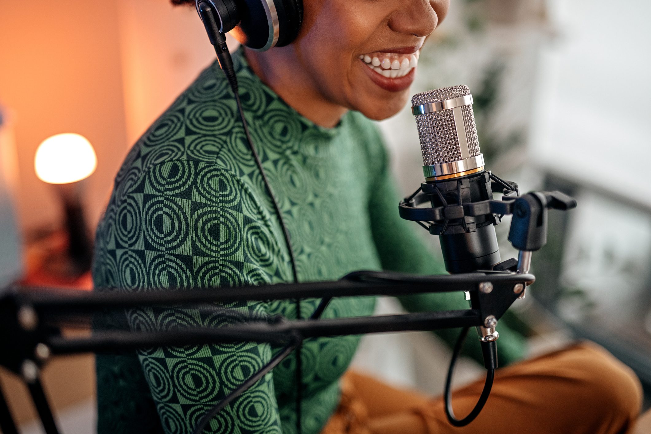 Microphone captures a person speaking, wearing headphones and a patterned green shirt, in a warmly lit indoor setting with a small lamp.