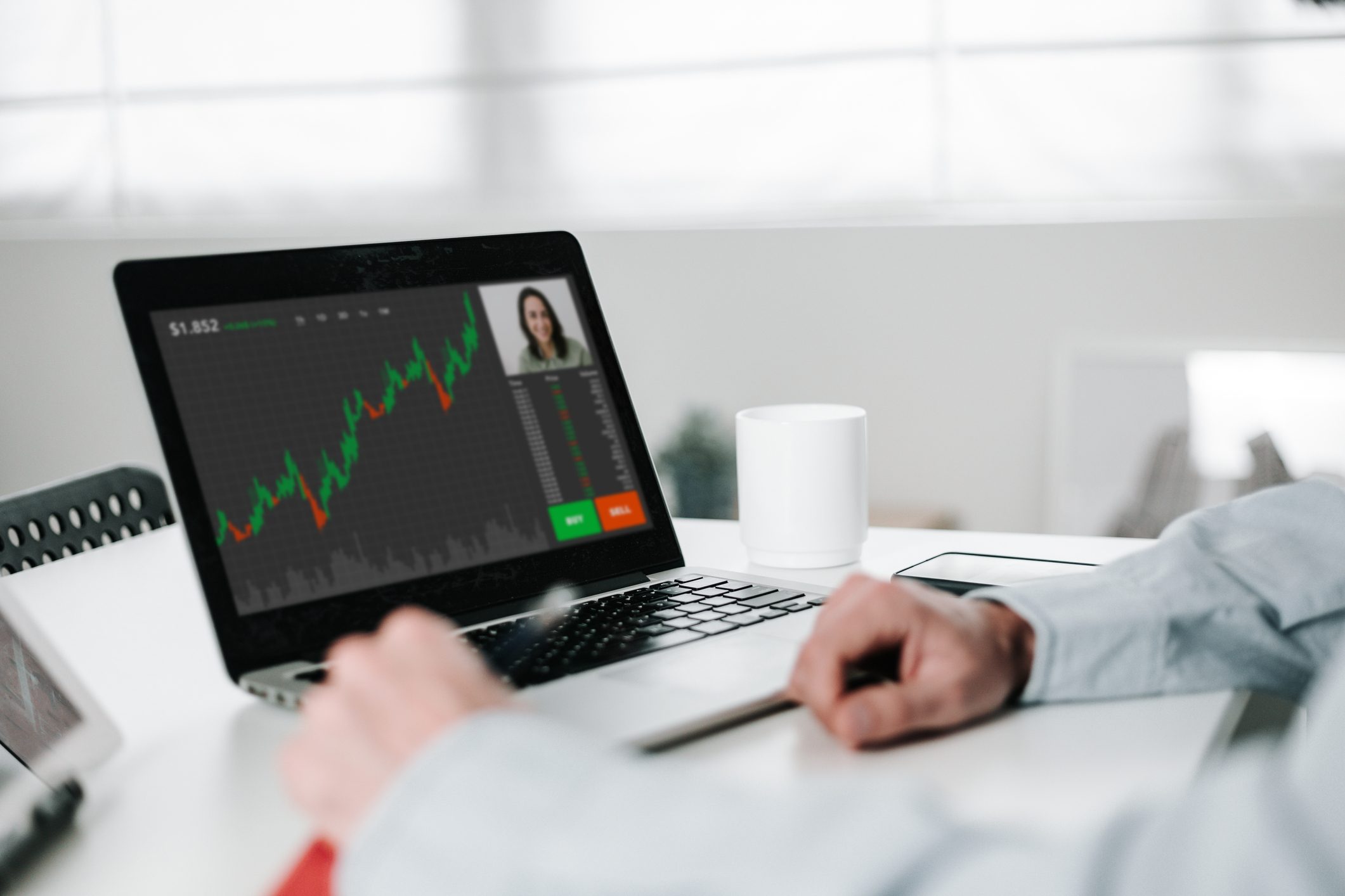 Laptop displays stock chart; person observes; on desk, a mug sits nearby in a bright, modern office.