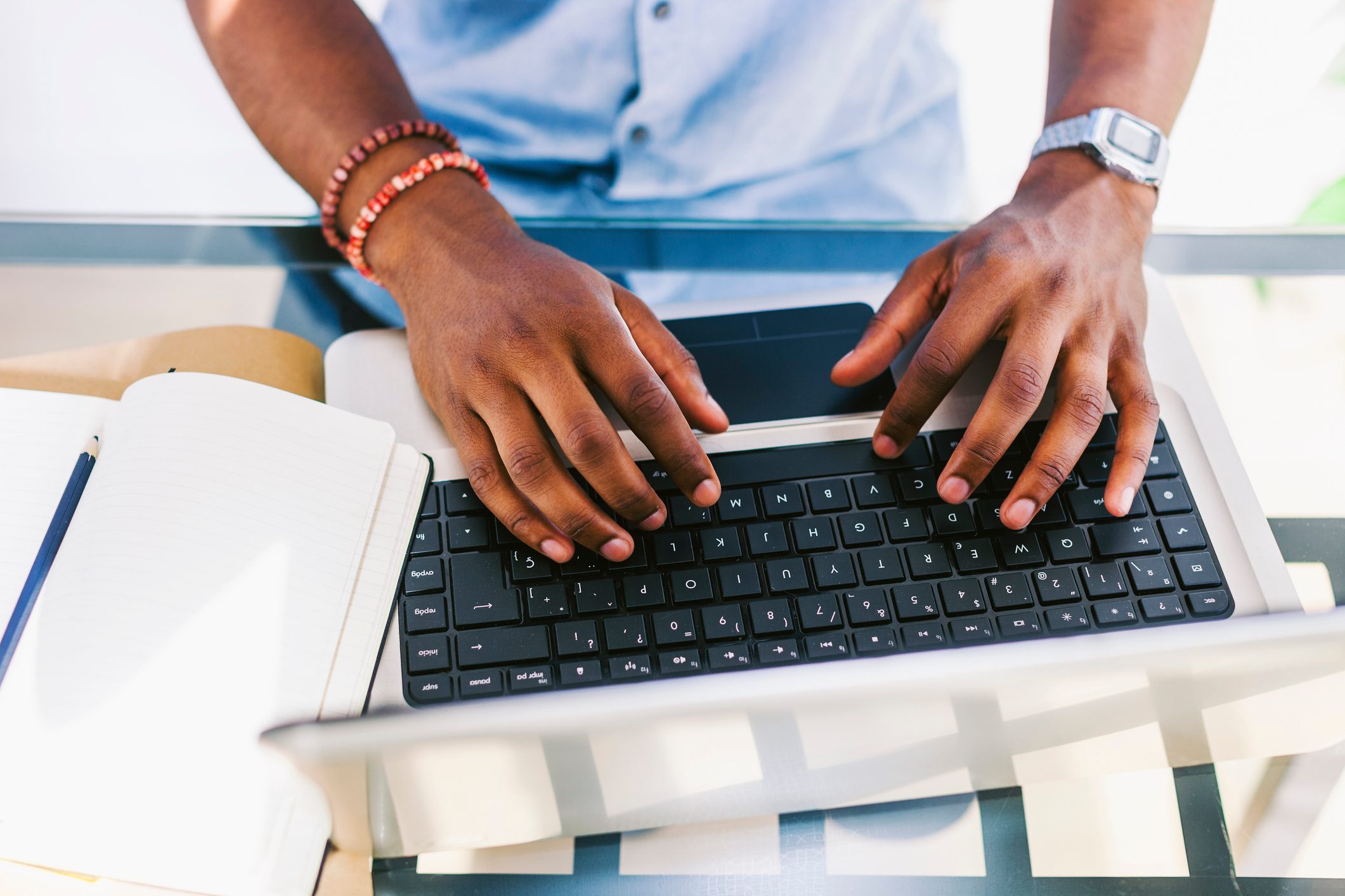 Hands typing on a laptop keyboard, with a notebook and pencil beside, set on a glass table.