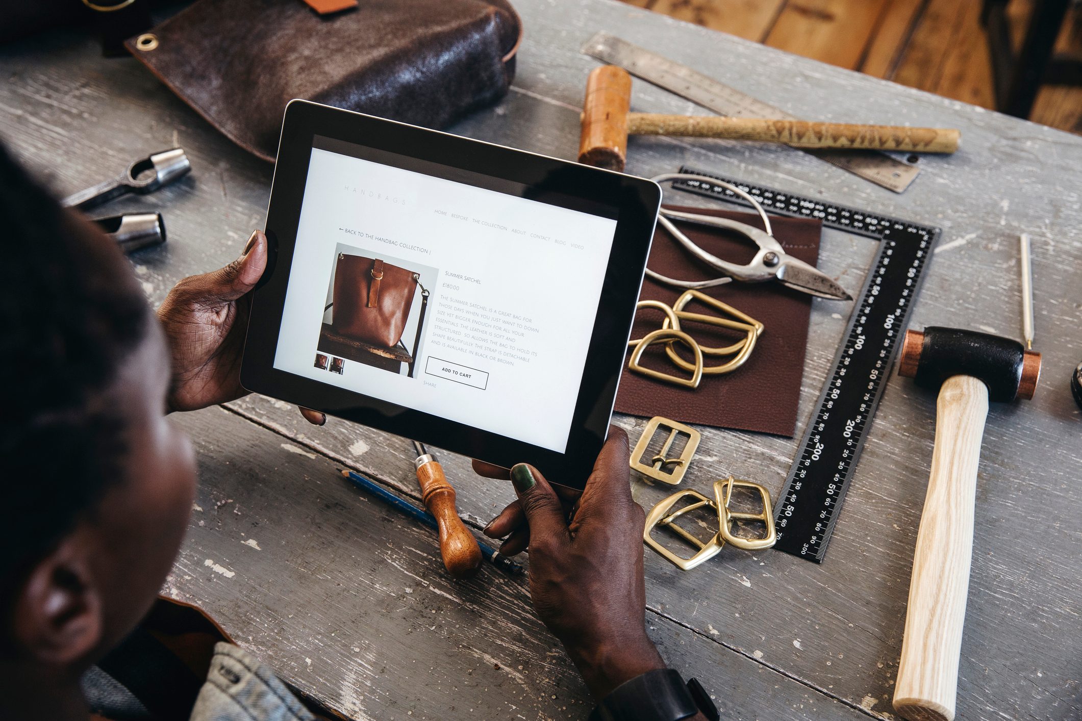 Tablet displays handbag information; person holds it in a workshop with tools and leather materials scattered on a wooden table.