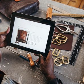 Tablet displays handbag information; person holds it in a workshop with tools and leather materials scattered on a wooden table.