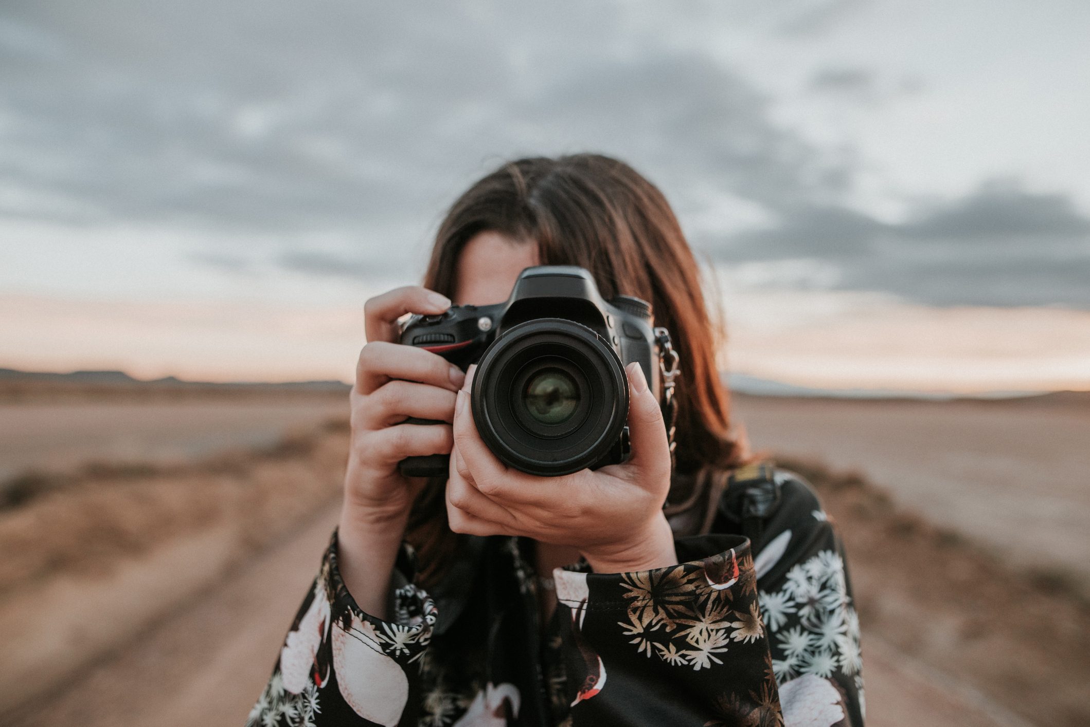 Camera held by a person taking a photo outdoors with a blurred landscape and cloudy sky in the background.