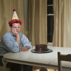 A person wearing a party hat sits alone, resting their face on their hand at a table with a chocolate cake, surrounded by curtains indoors.