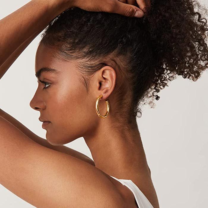 A person wears large gold hoop earrings, tilting their head with hands over a curly hairstyle, against a plain background.
