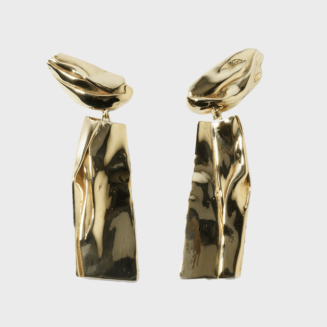 Gold earrings dangle, showing an abstract, sculptural design with reflective surface, set against a plain, light grey background.