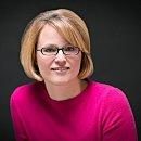 A person wearing glasses smiles slightly, seated against a dark background, wearing a bright pink sweater.