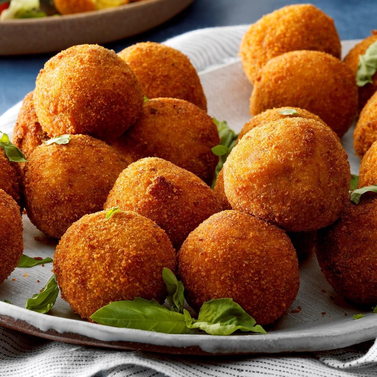 Golden, fried arancini sit on a white plate with fresh basil, creating an appetizing display on a textured cloth background.