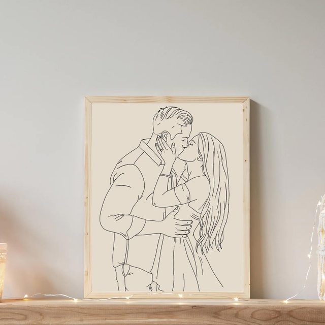 Framed line drawing depicts a couple kissing, placed on a wooden shelf, surrounded by soft string lights.