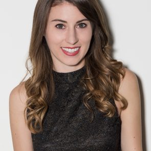 A person is smiling at the camera, wearing a patterned sleeveless top, with wavy hair in a neutral indoor setting.