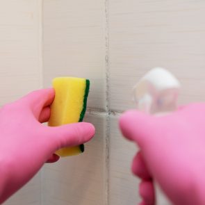 Hands in pink gloves scrubbing tiles with yellow sponge, applying cleaner, in a bathroom setting.