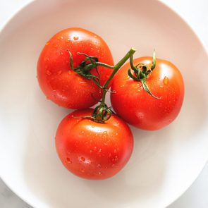 Three tomatoes resting on a white plate, covered with water droplets, creating a fresh appearance against a marble background.