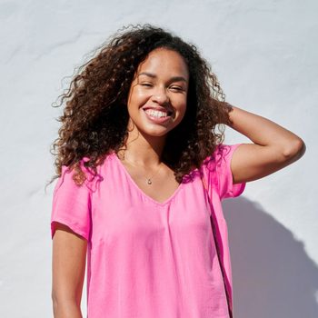 A person smiles with hand behind head wearing a pink shirt, standing against a plain white wall.