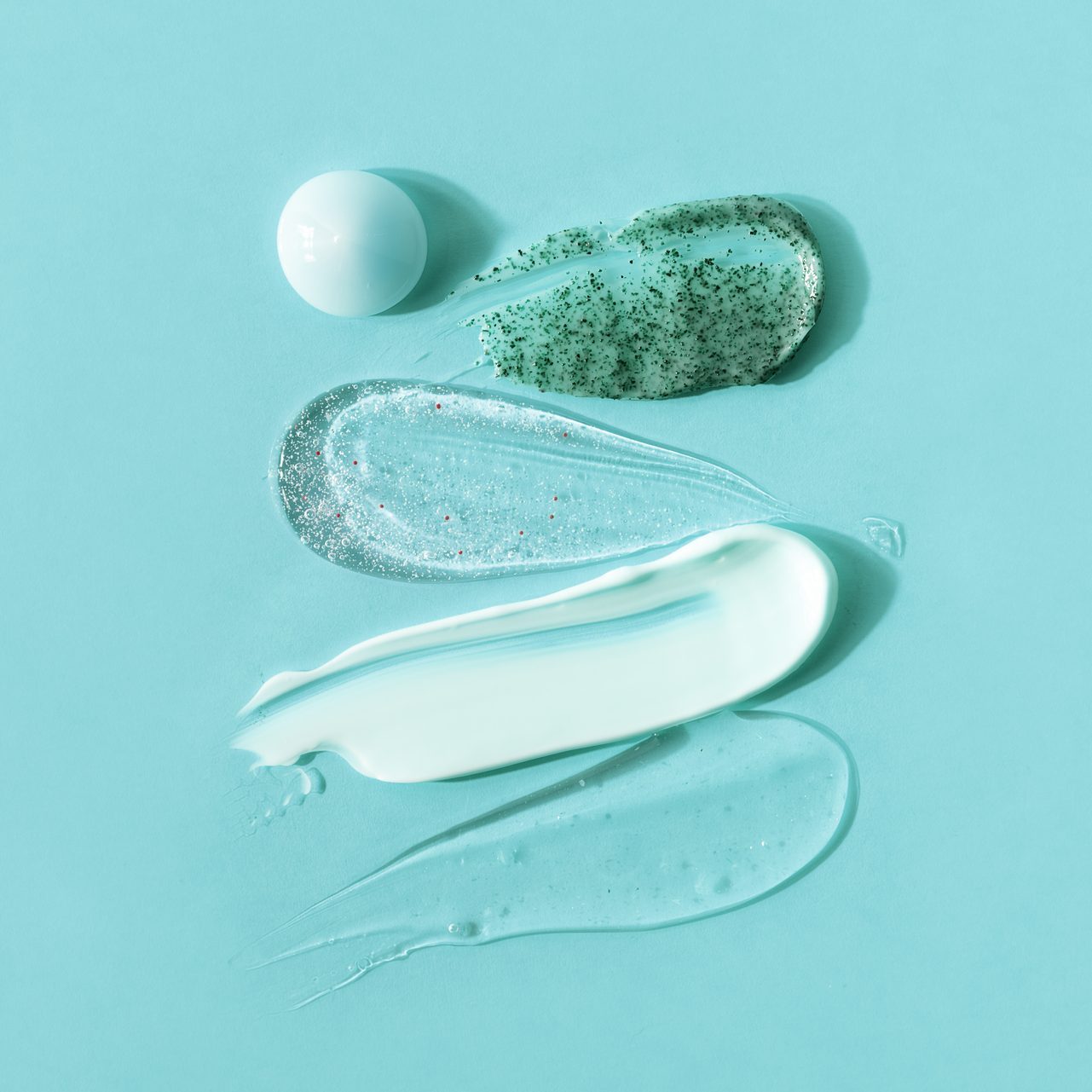 Swatches of skincare products spread on a light blue background; includes a translucent gel, glittery gel, cream, and a green exfoliant.