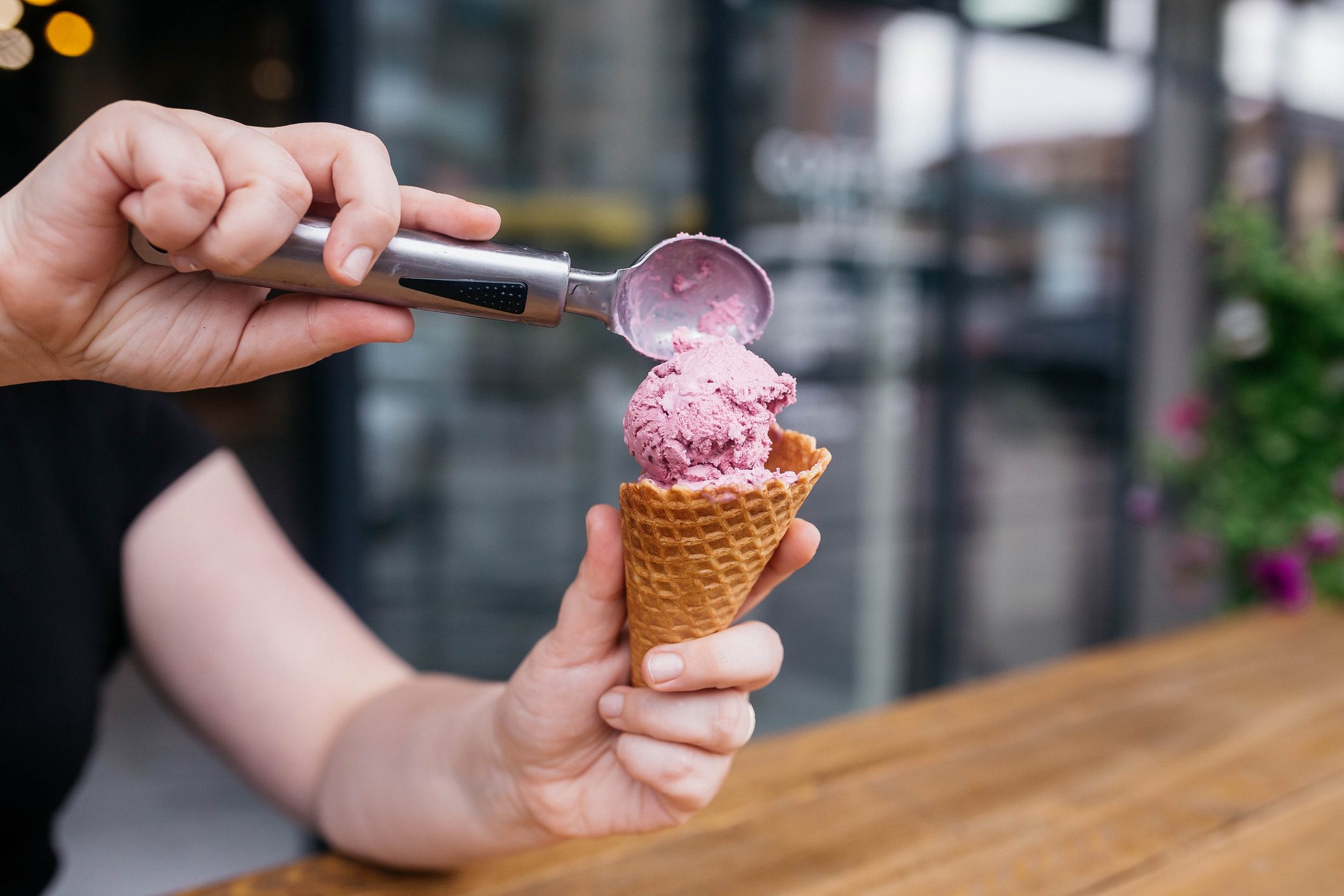 Scooped ice cream being placed on a cone by a person, set against a blurred outdoor cafÃ© background.