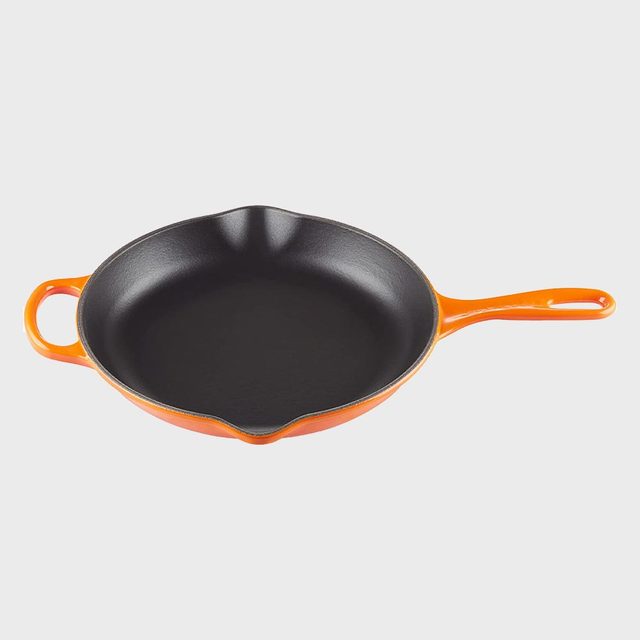 Orange cast iron skillet sits on a plain surface, featuring a long handle and helper handle.