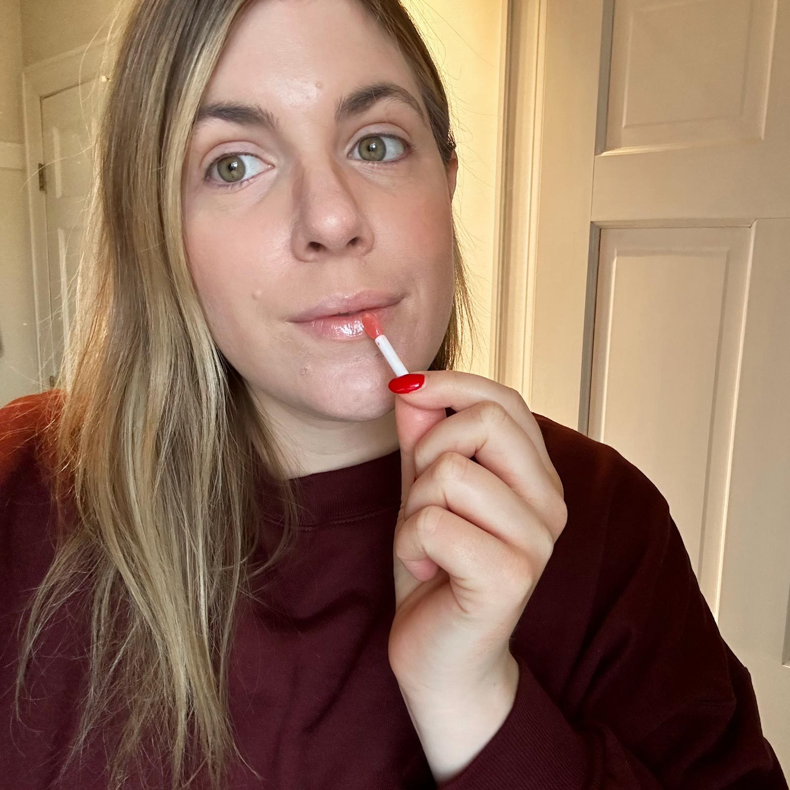 Person applies lip gloss, holding a small applicator, standing indoors with light-colored walls and door panels in the background.
