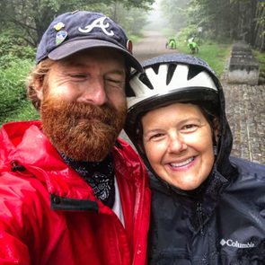 Two people smiling in rain gear, embracing on a misty forest trail. Bicycles and lush greenery are visible in the background.