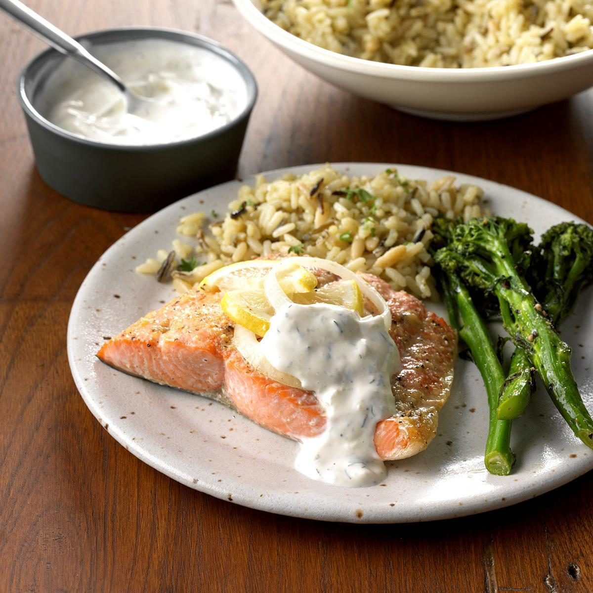 Salmon fillet topped with creamy sauce on a plate, accompanied by lemon slices, rice pilaf, and broccolini, set on a wooden table.
