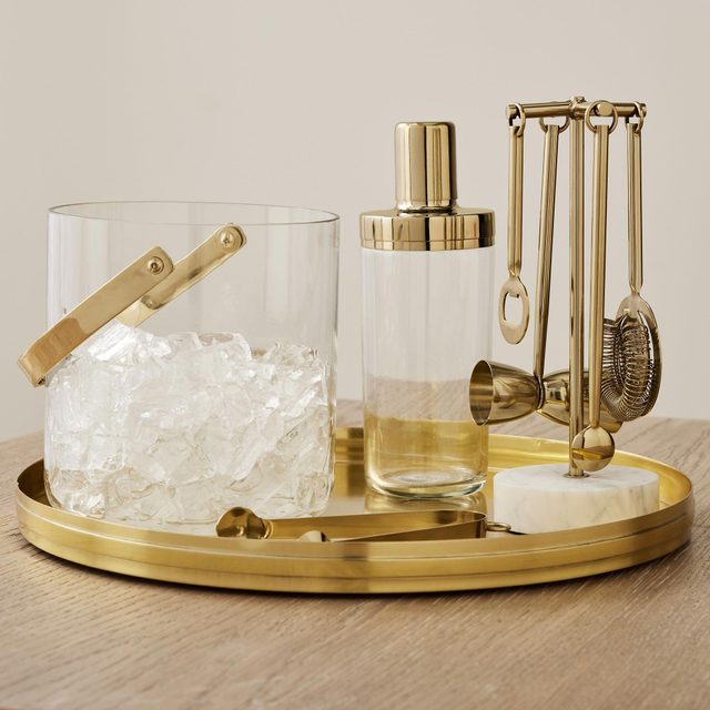 Ice bucket holds ice, alongside a cocktail shaker and bar tools, placed on a gold tray on a wooden table.