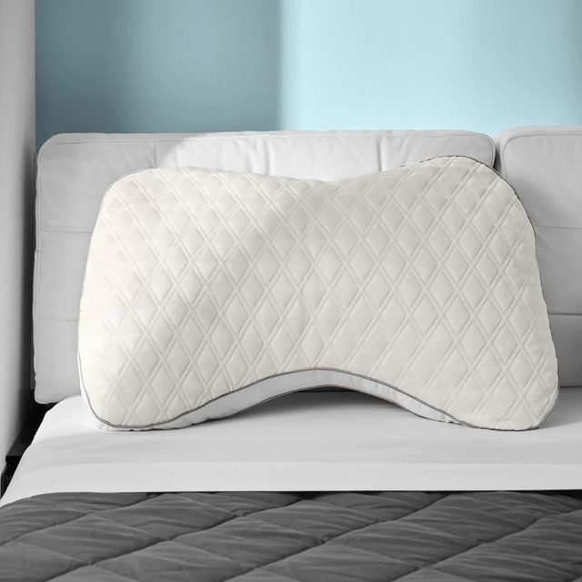 Curved pillow sits on a white bed, supported by a light blue wall, with a dark quilted blanket below.