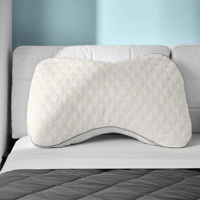Curved pillow sits on a white bed, supported by a light blue wall, with a dark quilted blanket below.