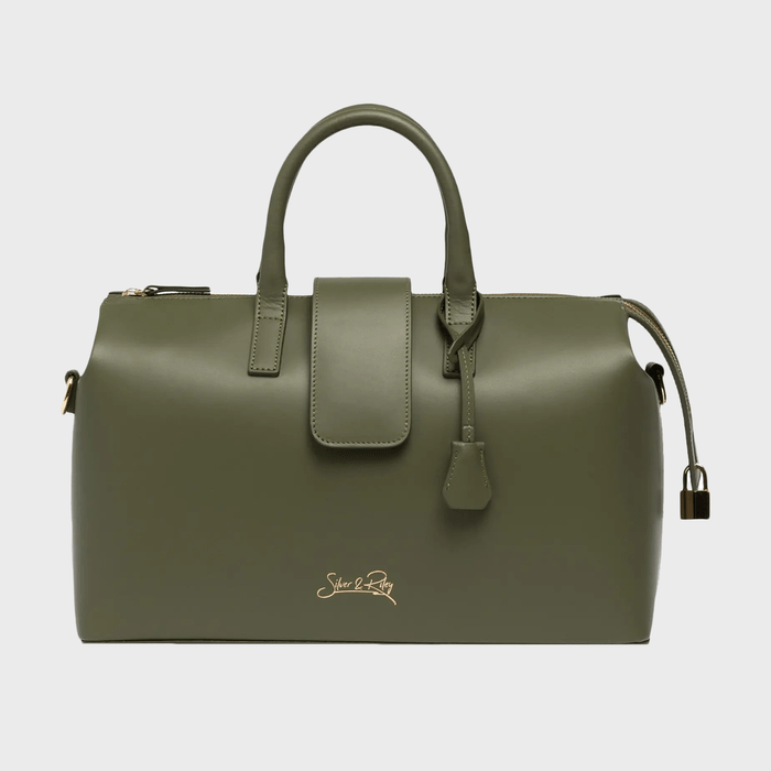 Green handbag resting against a plain background; features a central zipper, two handles, and a designer logo at the bottom.
