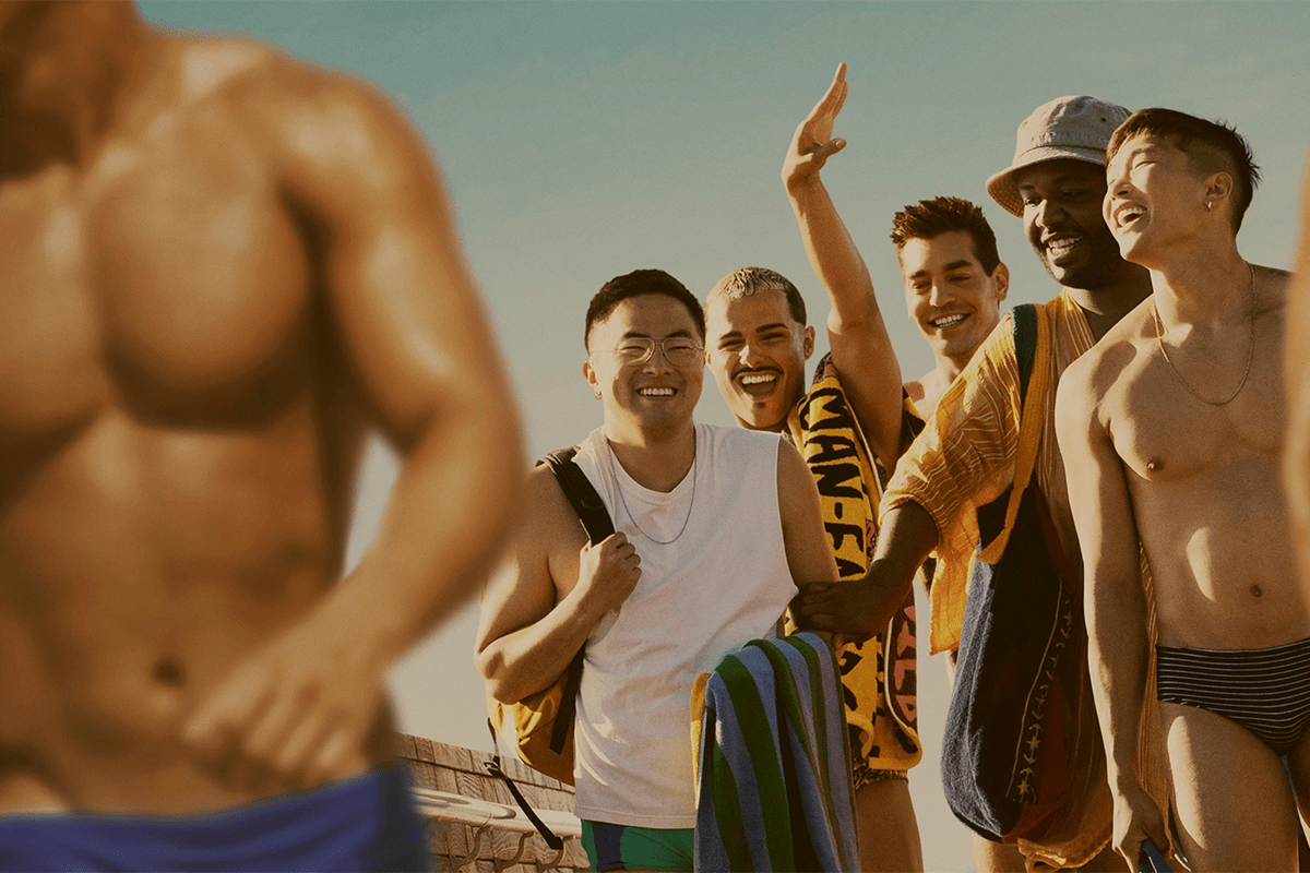 Group of people smiling, holding towels on a sunny beach. One person is pointing upwards. Blurred muscular figure in the foreground.