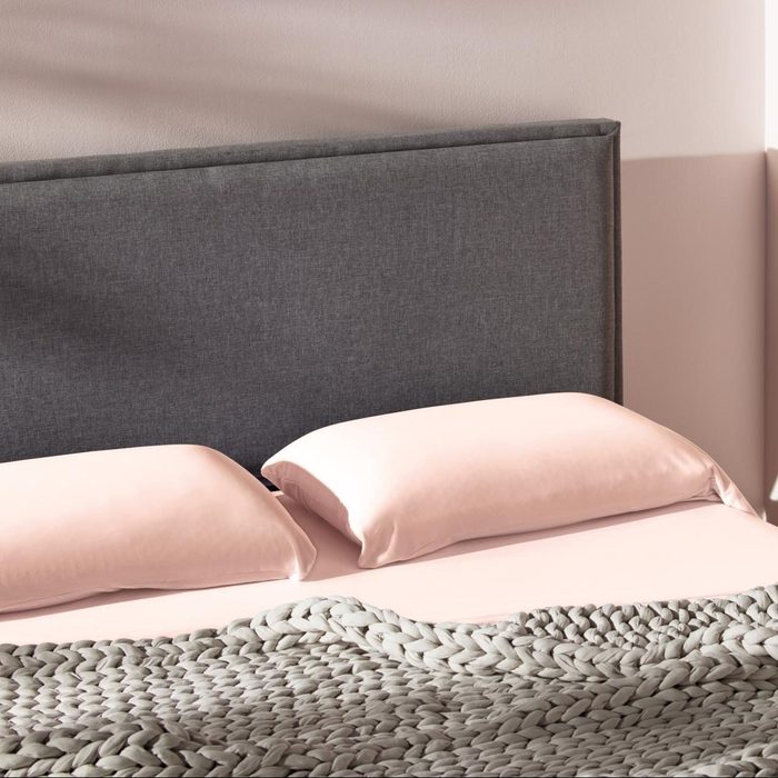 Two pink pillows rest on a bed with a gray knit blanket, positioned against a simple gray headboard in a softly lit room.